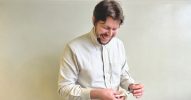 Mann in einem beigefarbenen Hemd steht vor der beigefarbenen Wand und putzt seine Brille.