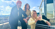 Ein Frau sitzt mit einem Smartphone in einem Einkaufswagen und wird von zwei weiteren Personen geschoben.