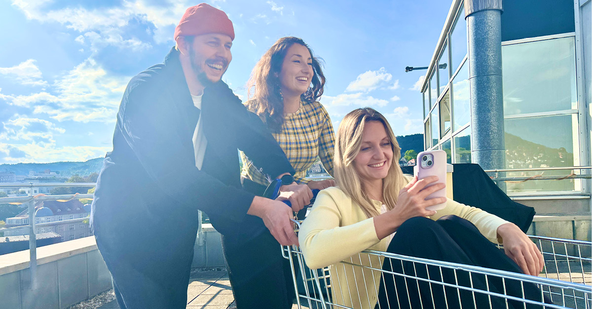 Ein Frau sitzt mit einem Smartphone in einem Einkaufswagen und wird von zwei weiteren Personen geschoben.
