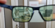 Eine Brille, die vor eine verschlossene Tür gehalten wird, gewährt den Blick auf zwei Personen, die an einem Tisch an einem Produkt arbeiten.
