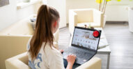 Eine Frau sitzt auf einem Sofa. Auf ihrem Schoß ist ein Laptop, auf dem ein Onlineshop geöffnet ist. An der Ecke des Laptops hängt ein großer roter Button.