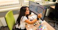 Eine Frau sitzt an einem Schreibtisch und arbeitet mit einem Pinsel an einem großen Monitor, auf dem eine Website oder digitale Oberfläche zu sehen ist.