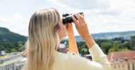 Frau blickt mit einem Fernglas über eine Stadt- und Landschaftskulisse.
