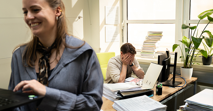 Mitarbeiter im B&uuml;ro wirkt gestresst am Schreibtisch, w&auml;hrend im Vordergrund eine Kollegin an einem Laptop arbeitet und l&auml;chelt. Sinnbild f&uuml;r fehlende Transparenz und steigende KI-Kosten im Unternehmen.