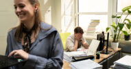 Mitarbeiter im Büro wirkt gestresst am Schreibtisch, während im Vordergrund eine Kollegin an einem Laptop arbeitet und lächelt. Sinnbild für fehlende Transparenz und steigende KI-Kosten im Unternehmen.